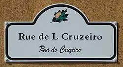 Photograph of a white street sign mounted on a tan wall reads in Mirandese “Rue de L Cruzeiro” with “Rua do Cruzeiro” written in Portuguese below in script, and a small leaf logo at the top.