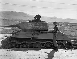 Soldiers stand around a destroyed tank with writing on it reading "Knocked out 20 July 1950 with the supervision of Maj. Gen. W.F. Dean"
