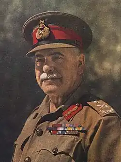 Head and shoulders colourised photograph of General Sir Thomas Blamey taken circa 1942. Blamey has a grey moustache and is wearing a peaked cap with scarlet cap band and general's bullion cap badge and an Australian Army khaki shirt to which are attached Australian general's embroidered rank slides, general's gorget patches in scarlet with gold bullion oakleaves and three ribbon bar rows for his various orders, decorations and medals.