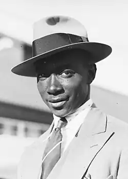 A head and shoulders black and white photograph man in a broad-brimmed hat and suit smiling at the camera.