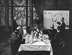 A black and white film still. A group of men sit around a dining table in the center. To the right, a man stands by and gestures at a large drawing of a dinosaur.