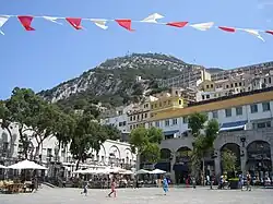 A broad, tree-lined sguare with shops and cafés on the perimeter and tiers of buildings on a slope leading up to the Rock of Gibraltar in the background