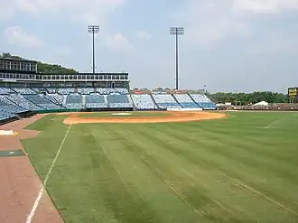 A view from the right field line of the seating bowl at Greer. Blue seats stretch from the right field wall, behind home plate, and beyond the third base dugout.