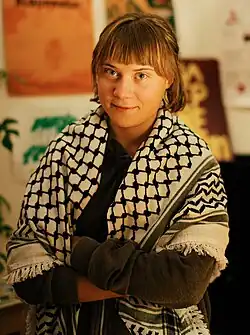 Portrait of Greta Thunberg. She is a young white woman with long blonde hair. She is wearing a black hoodie and a white keffiyeh around her.