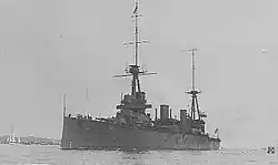 Photograph of a large warship. Along the bottom edge of the photograph, the phrases "HMAS AUSTRALIA" and "BUILT FOR THE AUSTRALIAN NAVY" are written in white. A sailboat and distant land can be seen in the left part of the photo.