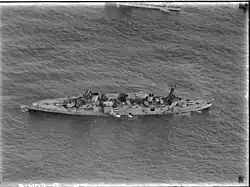 An aerial photograph of a large, World War I-era warship armed with four gun turrets, each with two guns, at sea. The ship is listing to port, but does not appear to be otherwise moving.