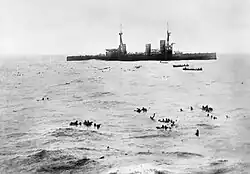 A large warship sits idly while several small boats pick up the numerous men swimming in the water.