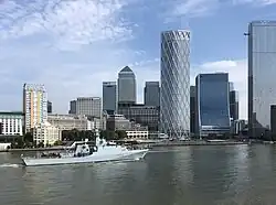 A grey naval patrol ship marked "P233" sails along a wide river in front of the Canary Wharf skyline, with modern glass skyscrapers and a partly cloudy blue sky in the background.