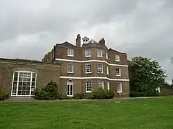 A three-storey brown brick building with a cupola, and a single storey extension on the left, the foreground is a green lawn