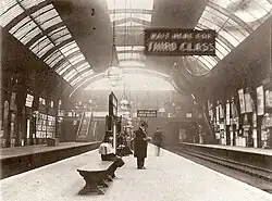 A black-and-white photograph of a railway station platform under a barrel roof. Several figures are visible, one standing wearing a top hat, a sign reads "WAIT HERE FOR THIRD CLASS".