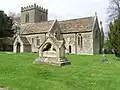 Gray stone building with small square tower at left hand end. In the foreground is grass with a small tiled memorial.