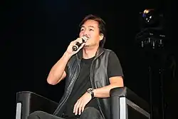 Low angle photograph of a mustached Japanese man in a black outfit sitting in a chair speaking into a microphone.