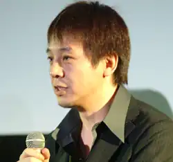 Portrait of a Japanese man with brown hair speaking into a microphone wearing a dark green suit jacket.