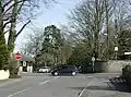 Street scene. Road junction with red stop sign. Two cars. The sides of the roads are stone walls with large trees showing above them.