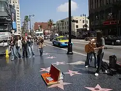 Two guitarists play on one side of a row of Walk of Fame stars, Hollywood Boulevard behind them, while pedestrians walk by on the other. Cars, including a blue and yellow taxi, and buildings can be seen in the bathroom.