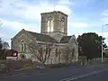 Stone building with arched windows and central square tower.In the foreground are trees and a road.