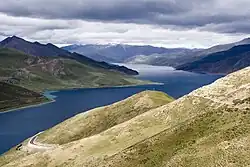 Yamdrok Tso, a large high-altitude lake in Tibet, surrounded by rolling green hills and distant mountains under a cloudy sky.