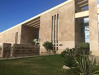 The entrance to the IRCAM (Institut Royal de la Culture Amazighe) building in Rabat. Two women are standing in front of a large, metal plaque with the word "IRCAM" written on it in large letters, and the organization's logo.