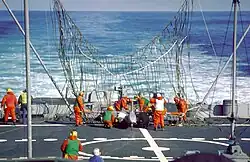 The rear deck of ship, with a large partially erect net visible near the center of the image. Many men in orange suits are working to free a white drone entangled in the net.