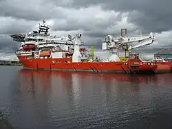 A red hulled ship seen from the port quarter at a low angle. It has heavy lifting gear on the quarterdeck and a helipad over the forecastle.
