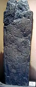 A shaft of stone with a badly damaged upper section. It bears the relief sculpture of a richly dressed human figure, standing and facing left. The head is missing.