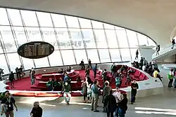 View of the headhouse's interior, facing the tarmac. There are people mingling in a lounge in the background. A sign hangs on the slanted glass wall next to the lounge.