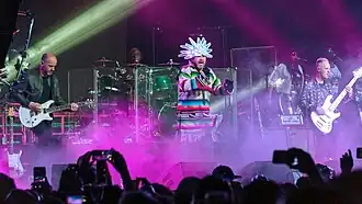 The band Jamiroquai performing on stage at Coachella 2018. At center is vocalist Jay Kay wearing a colourful Native-American shirt with frills around the waist and a white LED head-dress. Also present are a guitarist, a bassist, a drummer and two female backing vocalists. The band is engulfed in roughly waist-level stage fog coloured purple by stage lighting.