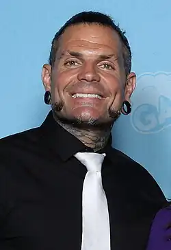 Man with short black hair and beard, with earrings and tattoed neck, smiling brightly at camera