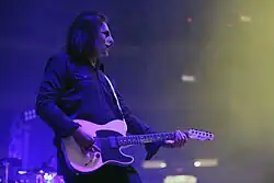 A man plays guitar on a major stage set wearing a black suit and a mask that resembles black and white makeup.