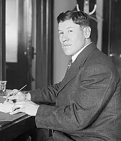 Black-and-white image of Jim Thorpe sitting down and writing. He is a Native American man with a Western haircut and wearing a dark suit.