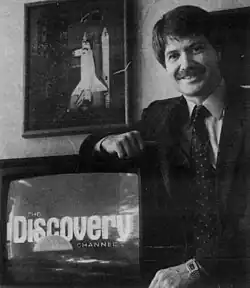 Black and white newspaper photo of John Hendricks, a light-skinned man with short dark hair and a mustache wearing a suit and tie, leaning on a CRT television displaying the logo of the Discovery Channel. On the wall behind him is a framed photo of a Space Shuttle on its launchpad