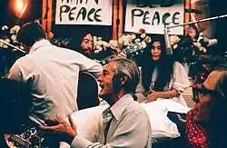 Lennon and Ono sit in front of flowers and placards bearing the word "peace". Lennon is only partly visible, and he holds an acoustic guitar. Ono wears a white dress, and there is a hanging microphone in front of her. In the foreground of the image are three men, one of them a guitarist facing away, and a woman.