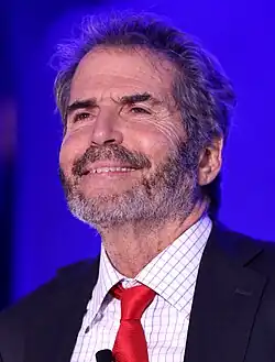Photograph of John Frank Stossel, an American television presenter, wearing a suit and red tie, smiling.