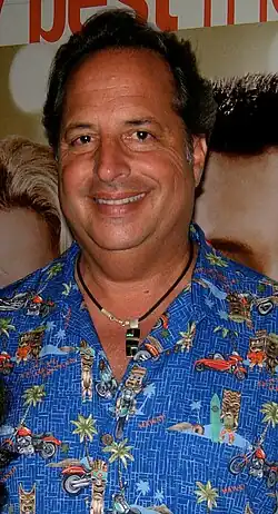 A black-haired man with a blue based shirt and a necklace looks straight at the camera smiling.