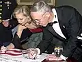 A young woman and an older man, both dressed in formal wear, leaning over to sign a poster.
