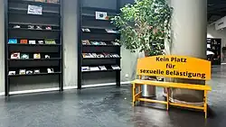 Library bookshelves in the background with a yellow two-seat wooden bench in the foreground. On its back, in black text, is written "Kein Platz für sexuelle Belästigung".