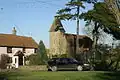 Stone church with square tower in the background partially obscured by trees. To the left is a pink painted house with red roof and in the foreground a car and grass area.