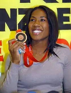 A Brazilian women in casual clothing holding a bronze medal up in her right hand