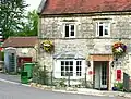 Stone building with old metal sign above the door saying Old Post Office, red post box set into the wall and to the left an old blue telephone box. The door is below the level of the road and separated by metal railings. Hanging baskets of flowers on the front of the building.