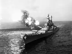 A black-and-white photograph depicting a large gunship sailing toward and slightly to the left of the camera. Guns of various size are visible on the ship, with smoke and flames visible from the turret No. 2 as the gun fires at an unseen target. The pressure from the gun fire has created a disturbance on the water surface