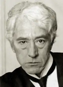 A white-haired man in his fifties, wearing a black jacket and tie and white shirt.