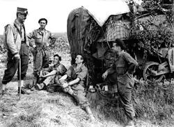 A man in uniform carrying a walking stick and wearing a kepi talks to four men in overalls wearing berets. A heavily camouflaged tank is in the background.