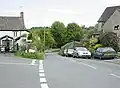 Street scene showing a road unction with parked cars. On the left and right are stone houses with trees and fields in the distance.