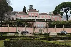 Photograph of Belém Palace. A large pale pink palace with tiled roofs sits behind a walled terrace and the Afonso de Albuquerque Square, with statues, trimmed hedges, and a few cars passing along the street in front.