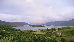 Green fields in the foreground with a lake beyond. There are wooded islets in the lake, which is skirted by low hills. Sunlight breaks through a clouded sky and illuminates a distant brown hillside.