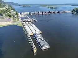 A low dam crosses a river. At one side: a rectangular pen holds barges.