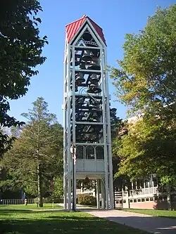 A three-dimensional vertical structure perhaps 70 feet (21 m) tall and 10 feet (3.0 m) wide by 10 feet (3.0 m) long is filled with tiers of large metal bells. The structure, standing in front of a large institutional building in a park-like setting, is supported by several metal legs. A sidewalk passes through the structure, which is open at the bottom to a height of perhaps 10 feet (3.0 m).