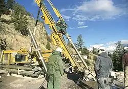 Two workers are looking at a piece of heavy machinery that is inserting a large drill downwards into the earth.