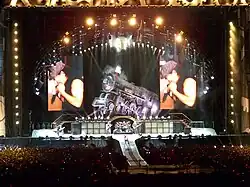 Distant shot of a concert stage. An arch with spotlights stands above a life-size locomotive and two big screens which display the male singer in left profile. Below and in front of the locomotive is musical equipment and four band members. At extreme left is a male playing guitar, he wears a school boy's uniform. Central stage has two guitarists with a drummer and his kit between them. Front stage has a catwalk with the singer turned to his left profile, he is cradling the microphone in his right hand and has his left fist near his face. He wears a cap, dark singlet and jeans.