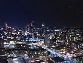 A skyline of a city against a blue sky and tall hills in the background. There are around nine skyscrapers distributed fairly evenly across the skyline with many cranes also visible. In the foreground there is a river with trees on both banks.